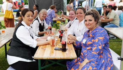 Mehrere Mitarbeitende sitzen an einer Bierbank mit Essen und Getränken und blicken in die Kamera beim Sommerfest 2026 auf dem Gesundheitscampus Volksdorf des Evangelischen Amalie Sieveking Krankenhauses, der Residenz am Wiesenkamp, des Feierabendhauses Volksdorf und der Albertinen Kindertagesstätte Volksdorf.