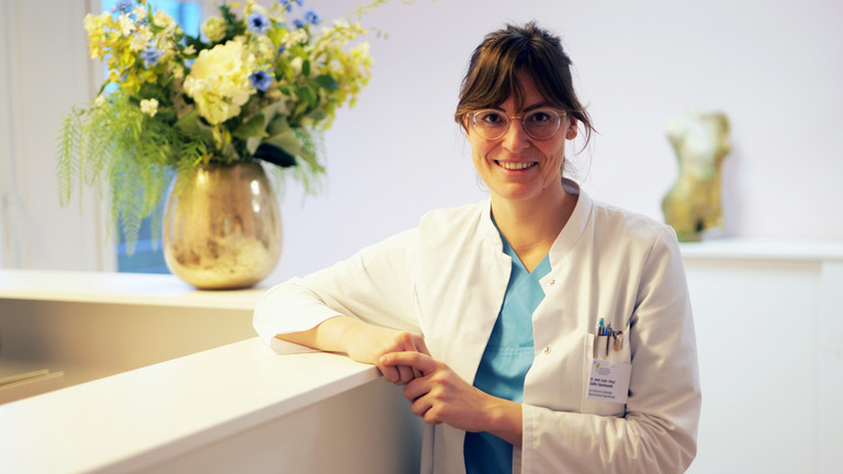 Dr. (univ. Pecs) Zsofia Danckwardt, Leitende Oberärztin Klinik für Gynäkologie und Geburtshilfe sowie Sektionsleiterin Urogynäkologie im Evangelischen Amalie Sieveking Krankenhaus in Hamburg, blickt angelehnt an einen Empfangstresen mit großem Blumenstrauß im Hintergrund in die Kamera