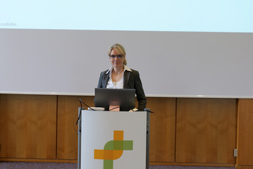 Eine Rednerin steht am Rednerpult mit einem Kreuzband-Logo der Immanuel Albertinen Diakonie vor einer Leinwand bei der Fachveranstaltung 11. Amalie Tag der Hausärztinnen und Hausärzte im Evangelischen Amalie Sieveking Krankenhaus in Hamburg-Volksdorf.