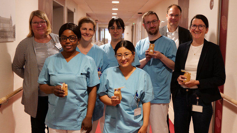 Mehrere Pflegekräfte in blauer Funktionskleidung halten kleine Präsente in den Händen und stehen mit Pflegedirektorin Maike Sellentin, ihrer Stellvertretrin Sabrina Oberle und Krankenhausgeschäftsführer Andreas Schneider in einem Krankenhausflur zum Internationalen Tag der Pflegenden 2026. 