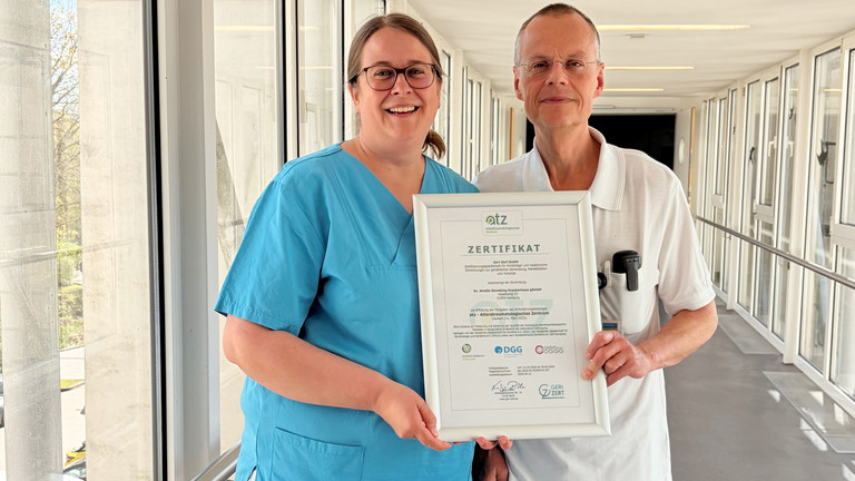 Dr. Saskia Otte, Chefärztin der Geriatrie und Physikalischen Medizin, und Dr. Olaf Pingen, Chefarzt der Orthopädie und Unfallchirurgie, stehen im Amalie Sieveking Krankenhaus in Hamburg-Volksdorf in einem hellen Durchgang und halten gemeinsam das Zertifikat für die erfolgreiche Zertifizierung des Krankenhauses als Alterstraumatologisches Zentrum in die Kamera. 