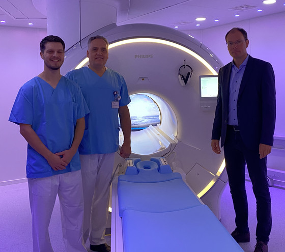 Pressefoto 2025-10-23: Evangelisches Amalie Sieveking Krankenhaus: Modernste MRT-Technologie für präzise Diagnostik und mehr Patientenkomfort Richard Grundherr, Prof. Christoph Weber und Andreas Schneider stehen in einem dunkellila illuminierten Raum vor dem neuen MRT-System im Immanuel Albertinen Zentrum für Radiologie am Standort Ev. Amalie Sieveking Krankenhaus in Hamburg und blicken in die Kamera
