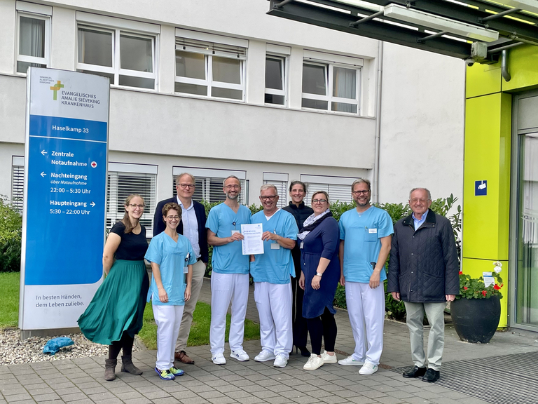 Mitarbeitende der Klinik für Anästhesiologie und Intensivmedizin zusammen mit Mitarbeitende der Ärztekammer Hamburg vor dem Krankenhaus mit dem Zertifikat in der Hand, Qualitätssicherung erfolgreich bestanden, Auszeichnung, Hamburg-Volksdorf