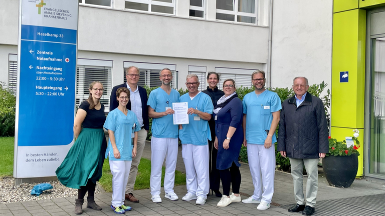 Mitarbeitende der Klinik für Anästhesiologie und Intensivmedizin zusammen mit Mitarbeitende der Ärztekammer Hamburg vor dem Krankenhaus mit dem Zertifikat in der Hand, Qualitätssicherung erfolgreich bestanden, Auszeichnung, Hamburg-Volksdorf