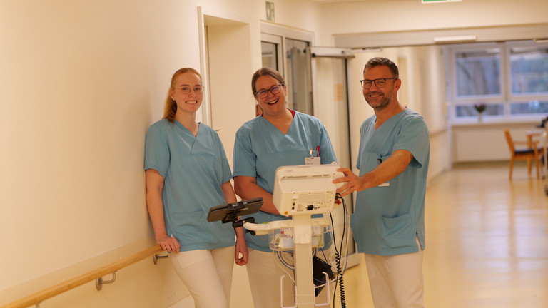 Drei Mitarbeitende des Amalie Alterstraumazentrum lachen in Arbeitskleidung vor einem Monitor in die Kamera