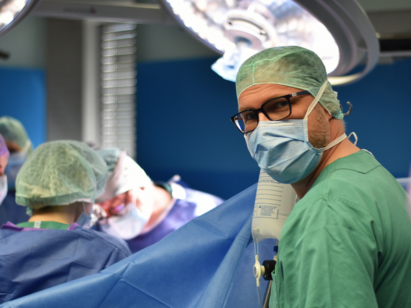 Mehrere medizinische Fachkräfte während einer Operation im OP-Saal, im Vordergrund schaut ein Arzt mit Mundschutz und OP-Kleidung in die Kamera