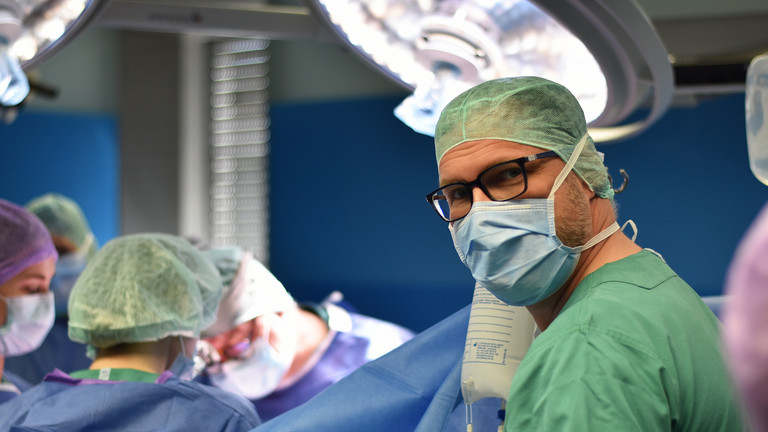 Mehrere medizinische Fachkräfte während einer Operation im OP-Saal, im Vordergrund schaut ein Arzt mit Mundschutz und OP-Kleidung in die Kamera