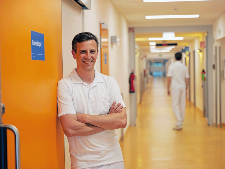Priv.-Doz. Dr. med. Johannes Kluwe, Chefarzt der Allgemeinen Inneren Medizin und Gastroenterologie am Evangelischen Amalie Sieveking Krankenhaus, steht in weißer Funktionskleidung an einer Schiebetür zur Endoskopie in einem Stationsflur und blickt in die Kamera.