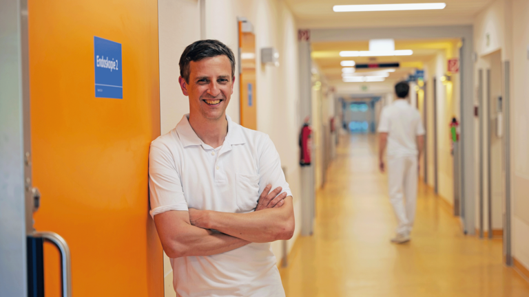 Priv.-Doz. Dr. med. Johannes Kluwe, Chefarzt der Allgemeinen Inneren Medizin und Gastroenterologie am Evangelischen Amalie Sieveking Krankenhaus, steht in weißer Funktionskleidung an einer Schiebetür zur Endoskopie in einem Stationsflur und blickt in die Kamera.