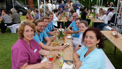 Mehrere Mitarbeitende sitzen in Funktionskleidung an einer mit Blumen geschmückten Bierbank mit Essensschalen und Getränken beim Sommerfest 2026 auf dem Gesundheitscampus Volksdorf des Evangelischen Amalie Sieveking Krankenhauses, der Residenz am Wiesenkamp, des Feierabendhauses Volksdorf und der Albertinen Kindertagesstätte Volksdorf. 