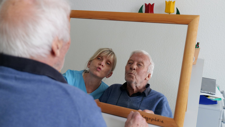 Ein Patient der Geriatrie & Physikalischen Medizin blickt gemeinsam mit einer Therapeutin in blauer Funktionskleidung in einen Spiegel während der logopädischen Therapie im Amalie Sieveking Krankenhaus in Hamburg-Volksdorf